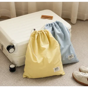 Pale yellow and light blue waterproof nylon drawstring bags with cord closures and small labels, beside a suitcase.