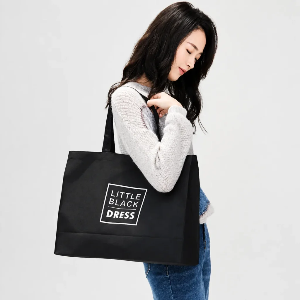 Black polyester Oxford carry bag with long black handles and white boxed lettering, carried on a woman's shoulder.